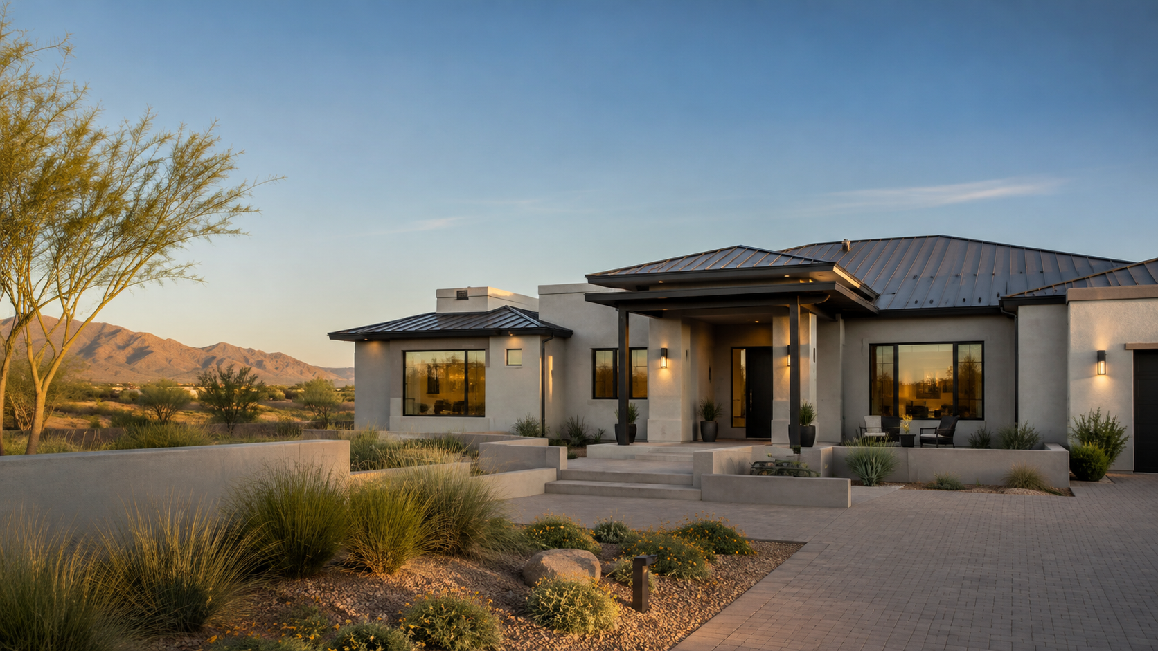 Modern desert-home façade with gray stucco, metal roof, and a covered entry, set against a sunset mountain backdrop.